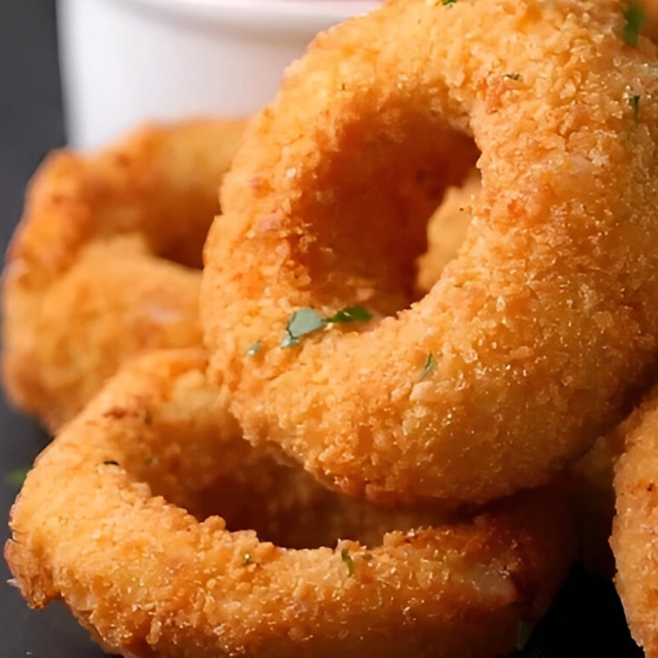 Close-up of golden-brown, crispy onion rings, lightly sprinkled with green herbs, on a dark surface.
